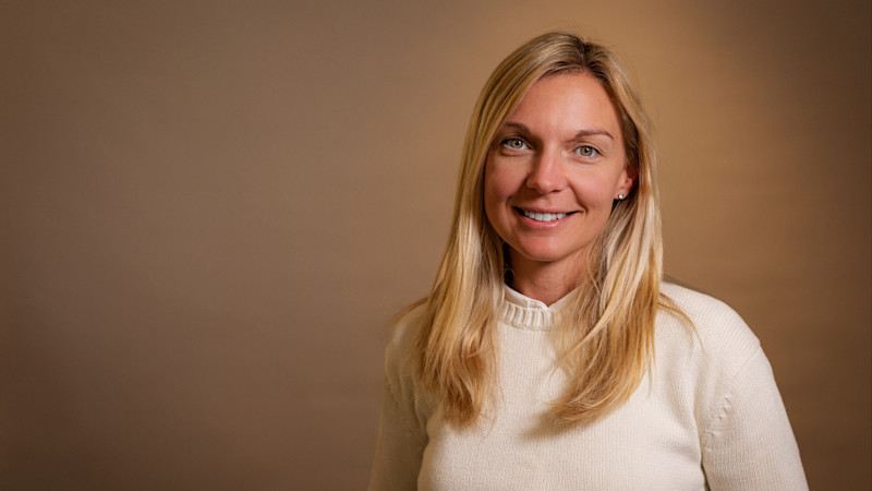 Porträtt av en leende person med långt blont hår i en vit tröja mot en brun bakgrund.
