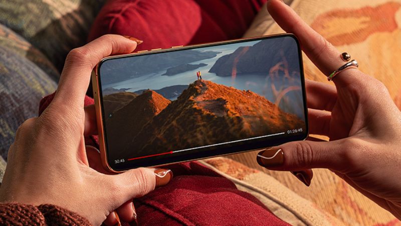 En person håller i en Motorola Edge 50 Neo och tittar på en video av ett bergslandskap med en person som står på toppen. Skärmen visar skarpa och levande färger. Personen har en brun stickad tröja och bär ringar på sina fingrar.