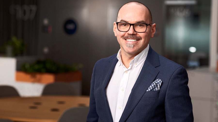 Porträttbild på Stefan Trampus, Executive Vice President för Tele2 Företag. Han har mustasch och glasögon och ler. Han är klädd i mörkblå kavaj och vit skjorta och har en blåvitrutig näsduk i bröstfickan. I bakgrunden syns ett kontor med mörkbruna väggar och möbler samt ett runt bord med stolar runt.