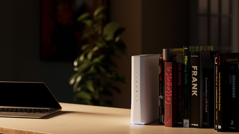 Ett modernt skrivbord med en vit trådlös router placerad mellan flera böcker, vilket får den att smälta in som en del av bokraden. På skrivbordet finns även en öppen laptop. I bakgrunden syns en grön växt och en tavla i dämpad belysning, vilket skapar en varm och hemtrevlig miljö.
