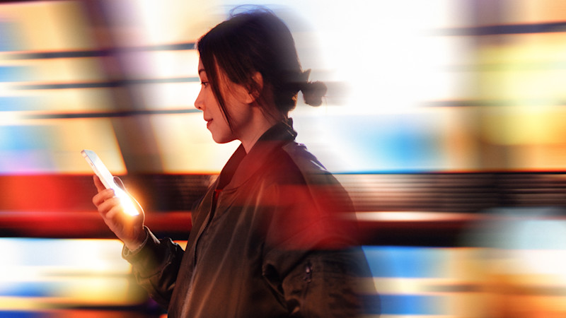 Profil av person med hårknut som tittar på lysande mobiltelefon mot suddig bakgrund med färgglada ljusstråk.