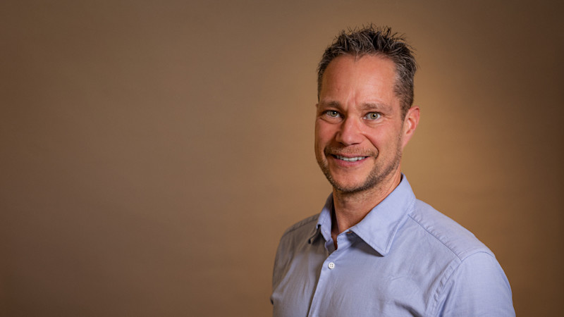 En leende person i ljusblå skjorta mot en varm brun bakgrund i ett professionellt porträtt.