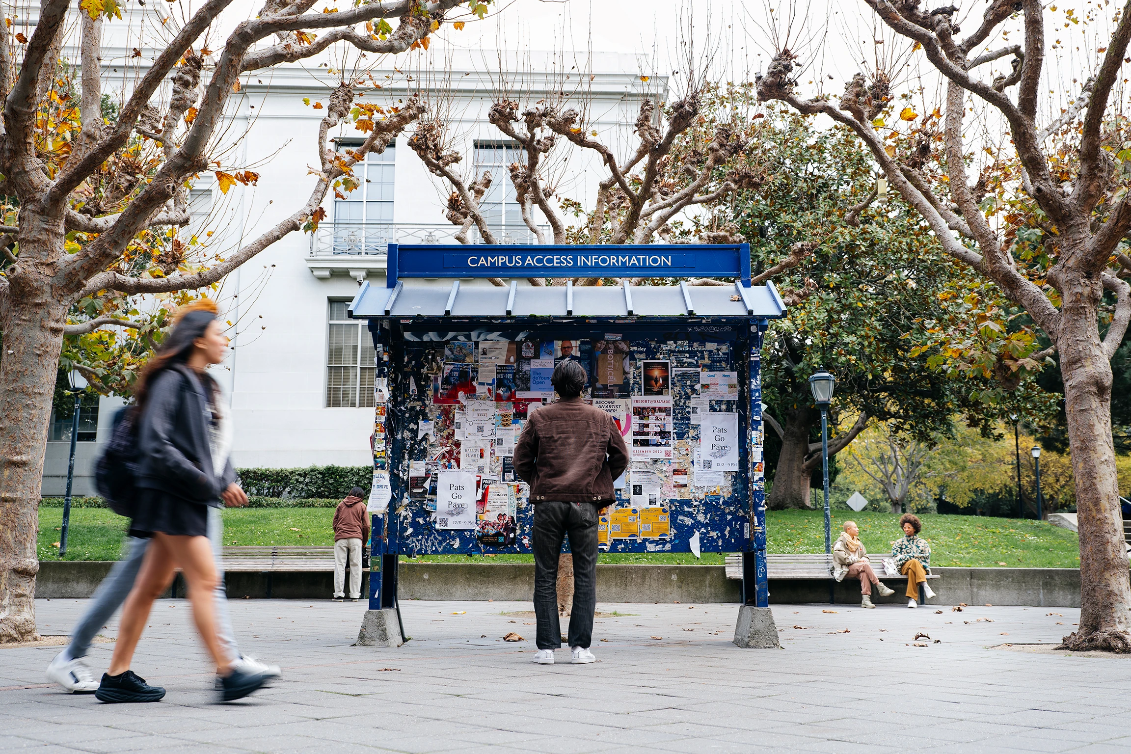 A man in a brown jacket stands in front of blue bulletin board that is covered with papers and advertisements University of California Berkely campus. Photo: Elena Zhukova for Mellon Foundation