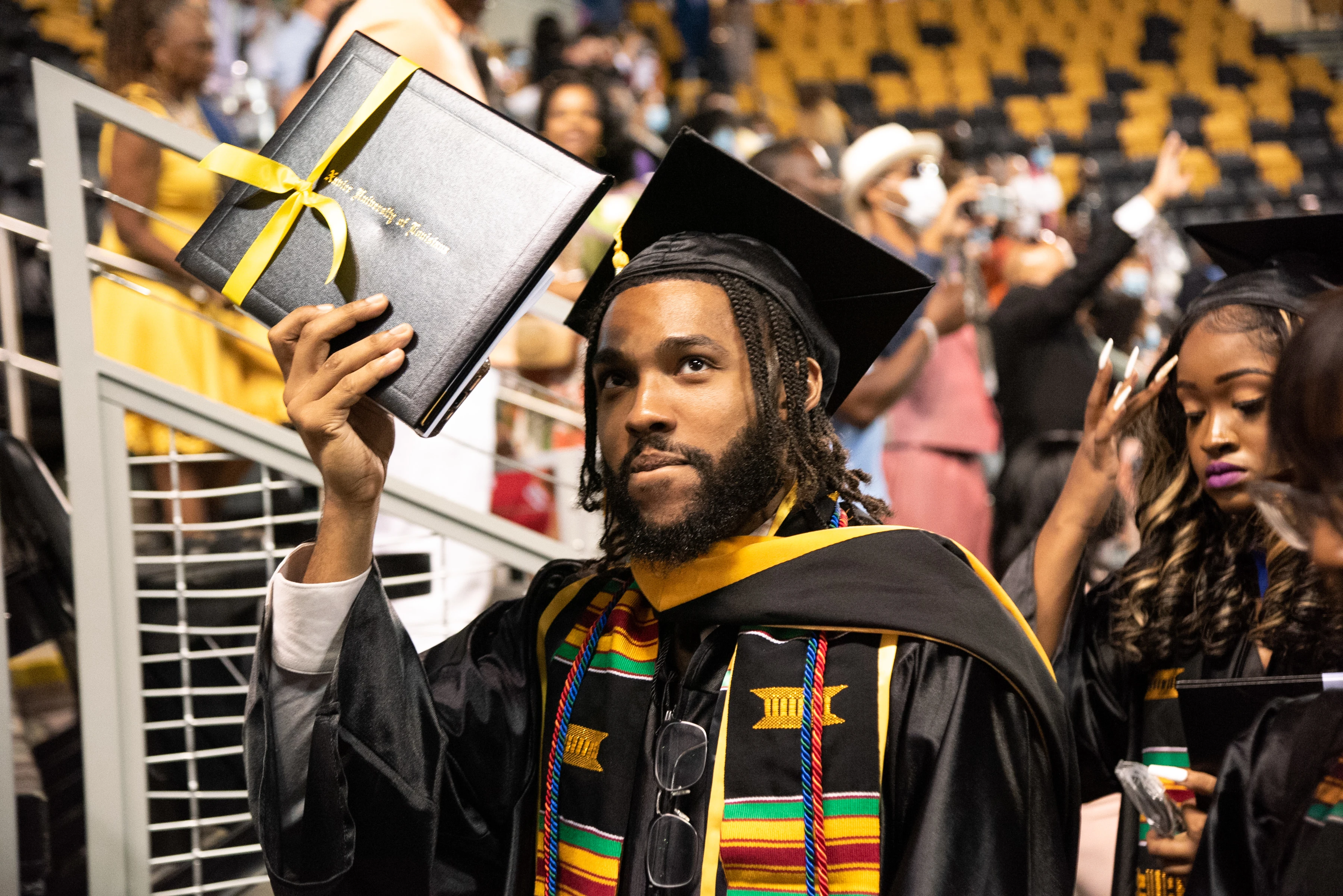 An African American man with a beard in a cap and gown holding up his diploma at a graduation ceremony