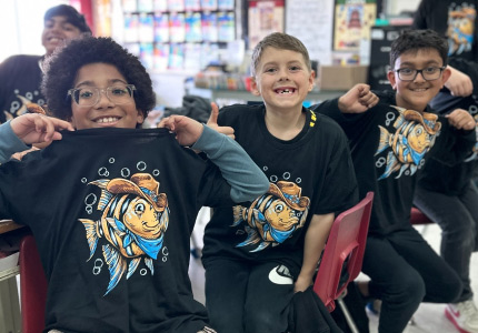 Boys sitting in a class room proudly wearing their black t-shirts featuring an angelfish wearing a cowboy hat.