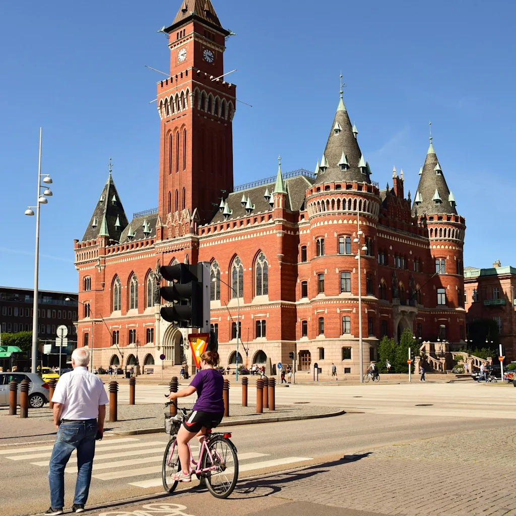 En storslått rød mursteinsbygning med klokketårn i Helsingborg, med folk som sykler og går på en solrik bygata.