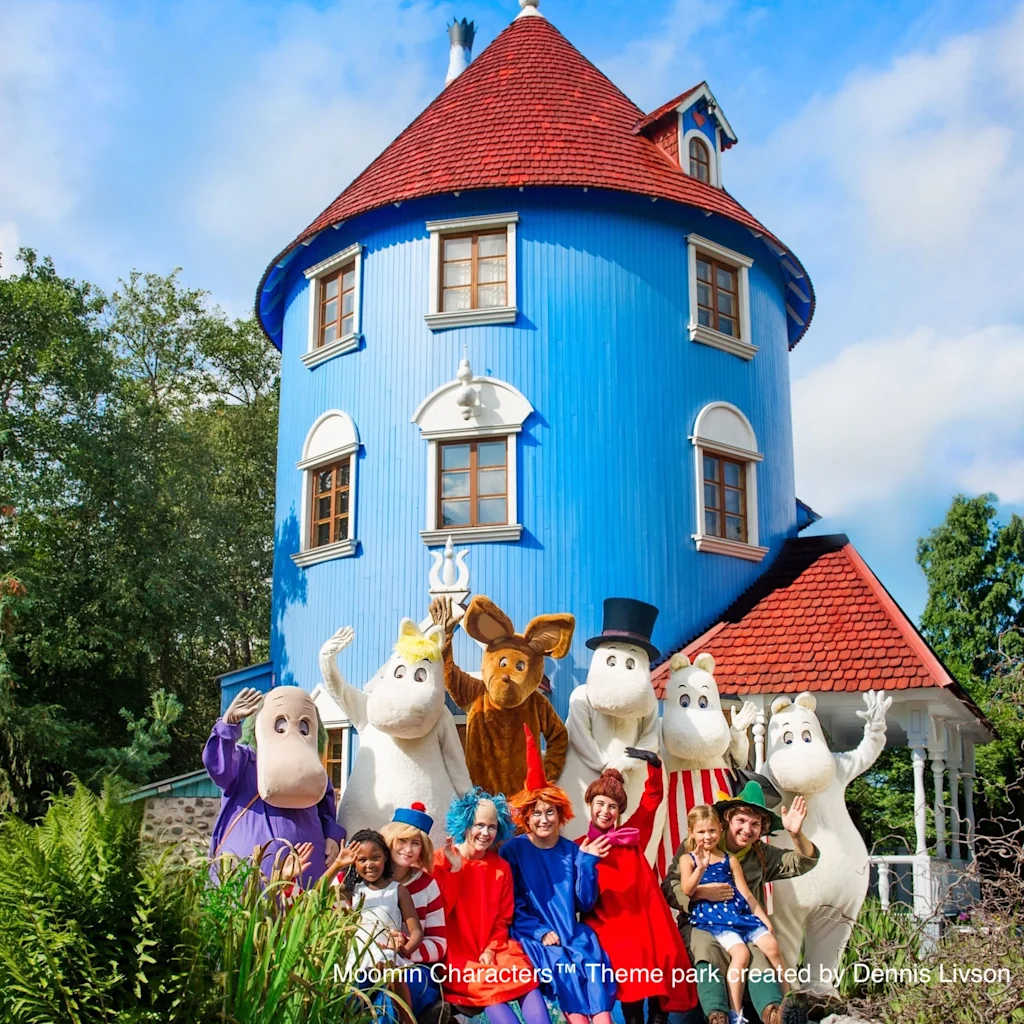 Ryhmä ihmisiä ja Muumi-hahmoja iloisesti vilkuttamassa erottuvan sinisen talon edessä, jossa on punainen katto.