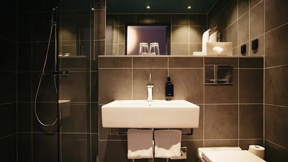 Et moderne badeværelse med håndvask, spejl, bruser og toilet i et mørkeflisebelagt rum med varmt lys.