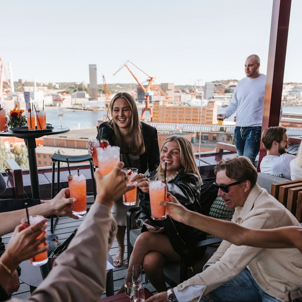 A group of friends enjoying cocktails and snacks on a rooftop terrace with a city view, toasting and laughing.