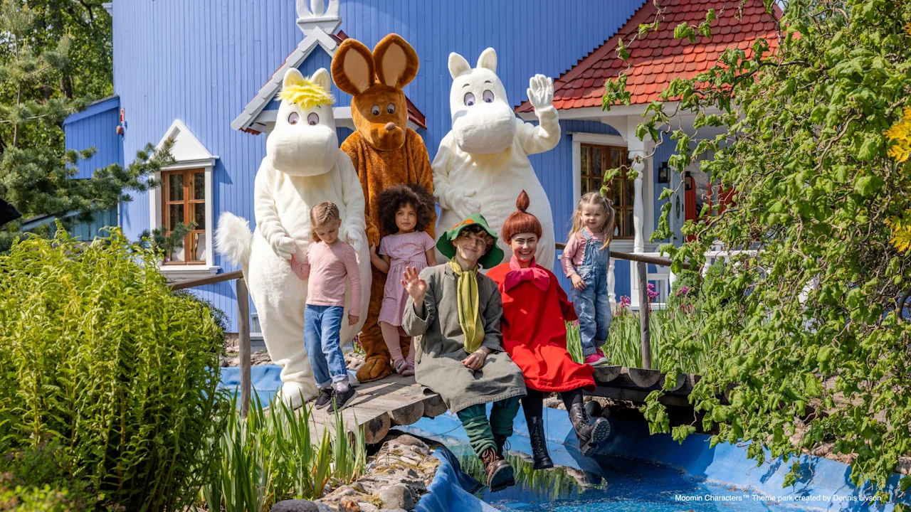 Lapset ja puvustetut hahmot poseeraavat iloisesti sillalla sinisen puron yli, sinisen talon ja vehreän luonnon edessä.