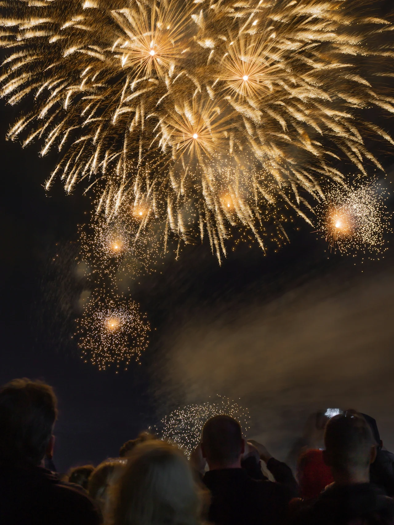 Fireworks explode in a night sky, illuminating the dark backdrop with golden light. A crowd of onlookers observes the spectacle, capturing the moment with cameras and phones.