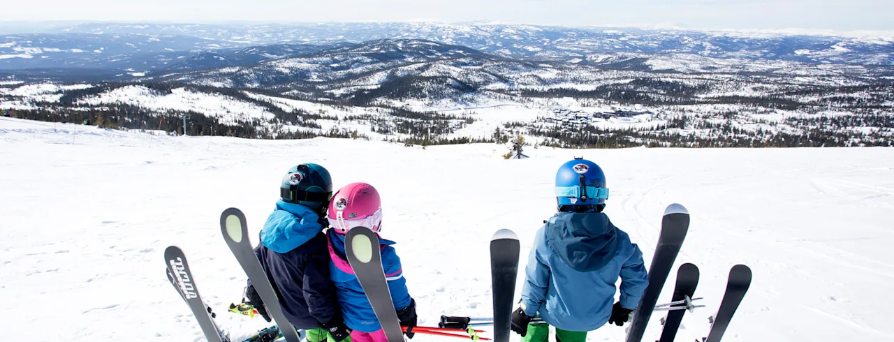 Three children in ski gear looking out at a vast, snowy mountain landscape on a bright winter day.
