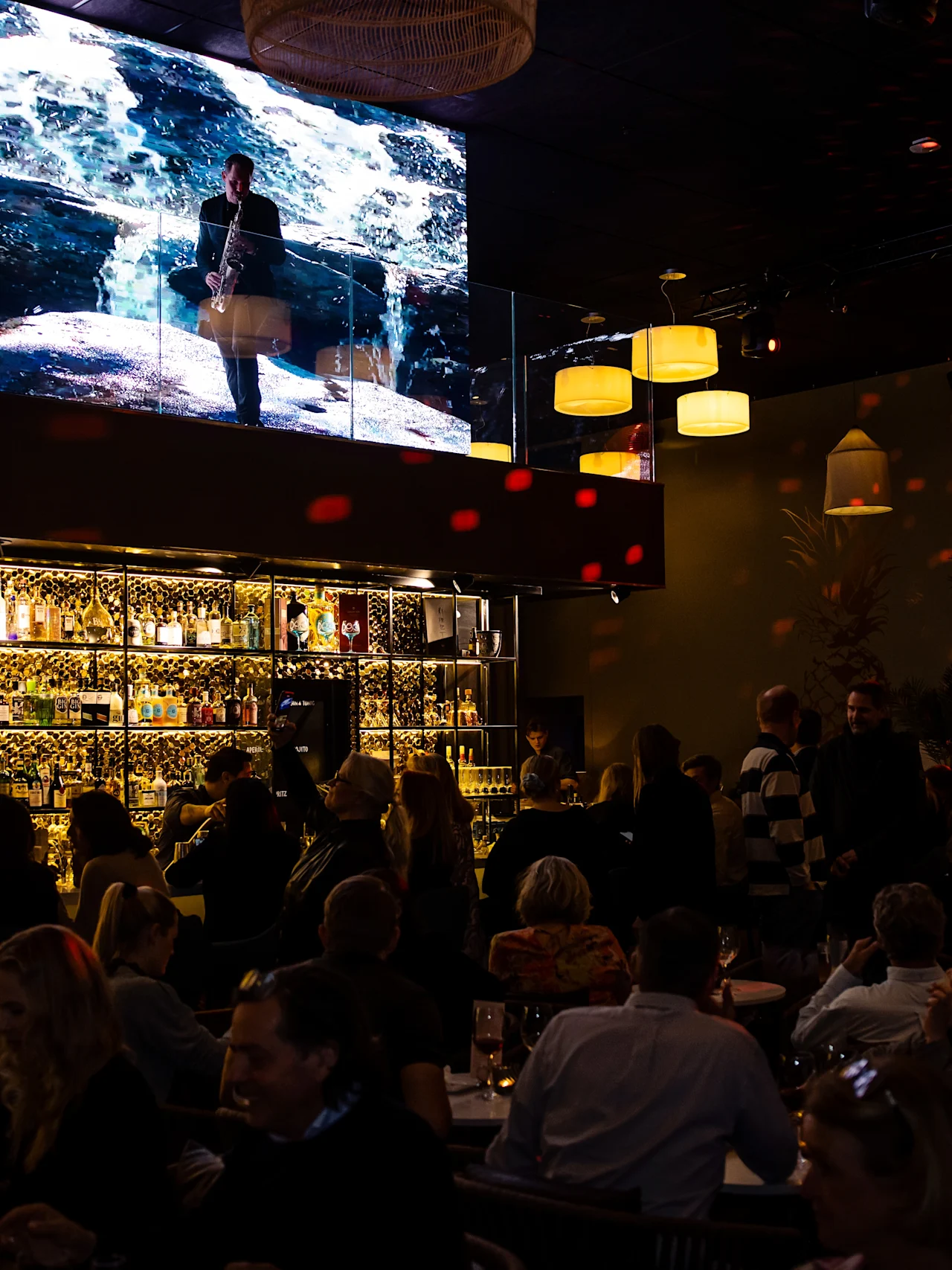 A bar is crowded with people socializing under warm lights. Above, a large screen displays ocean imagery. Shelves behind the bar are filled with bottles, creating a lively atmosphere.