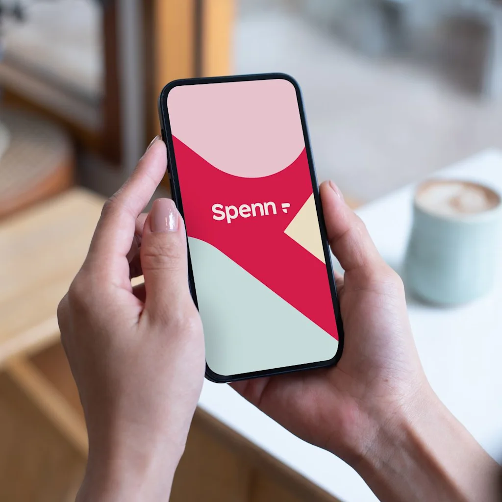 En person holder en smartphone med en app-grænseflade, der viser teksten "Spenn" over et rødt og beige design, ved et bord med en kaffekop.