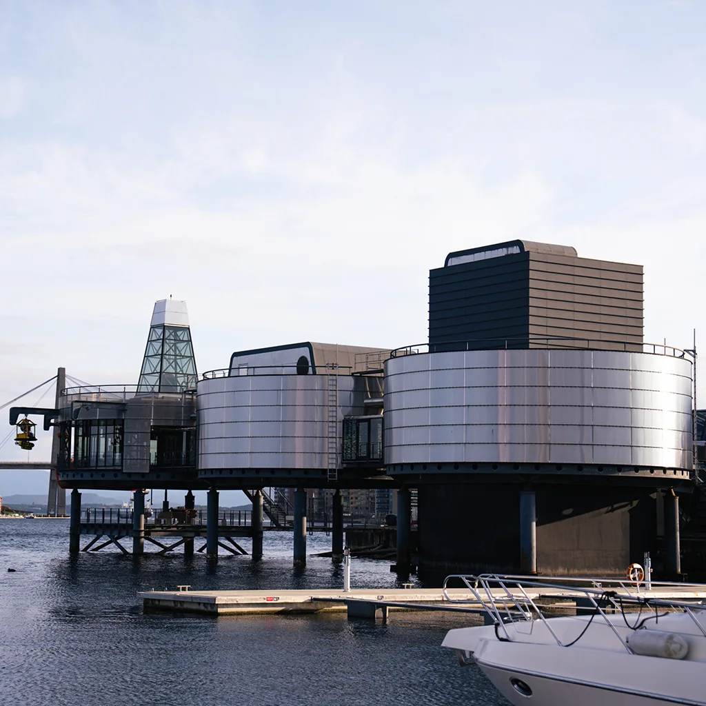 Nykyaikainen rakennus ulottuu tyynen veden ylle, taustalla kaukainen silta, rannikon koteja ja vene etualalla.