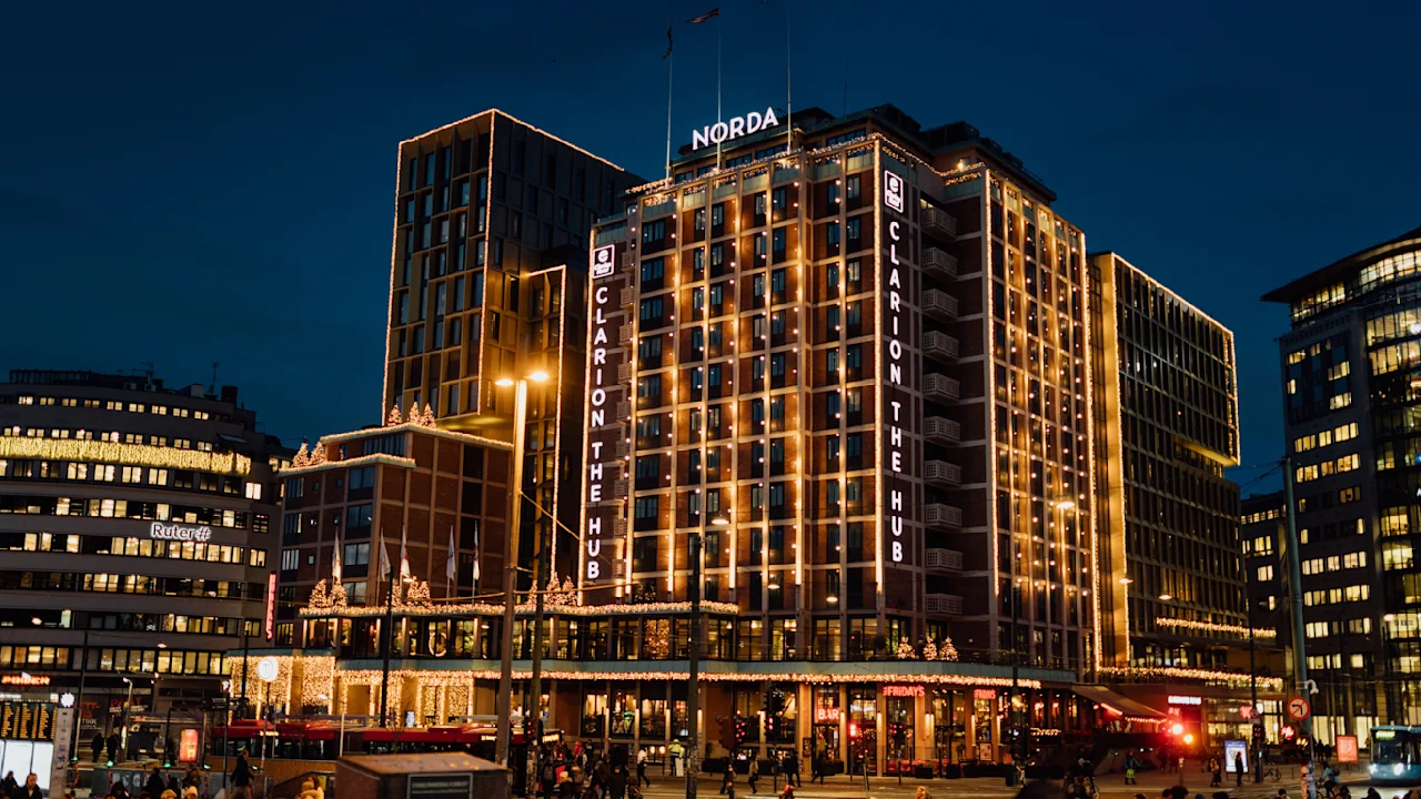 A multi-story building, illuminated with lights, displaying the texts "CLARION THE HUB" and "NORDA" at night. It is surrounded by people walking in a busy urban area.