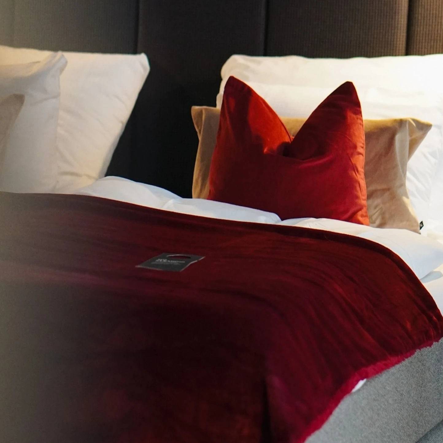 A comfortable bed with a dark upholstered headboard, soft white pillows, a vibrant red accent pillow, and a luxurious red throw.