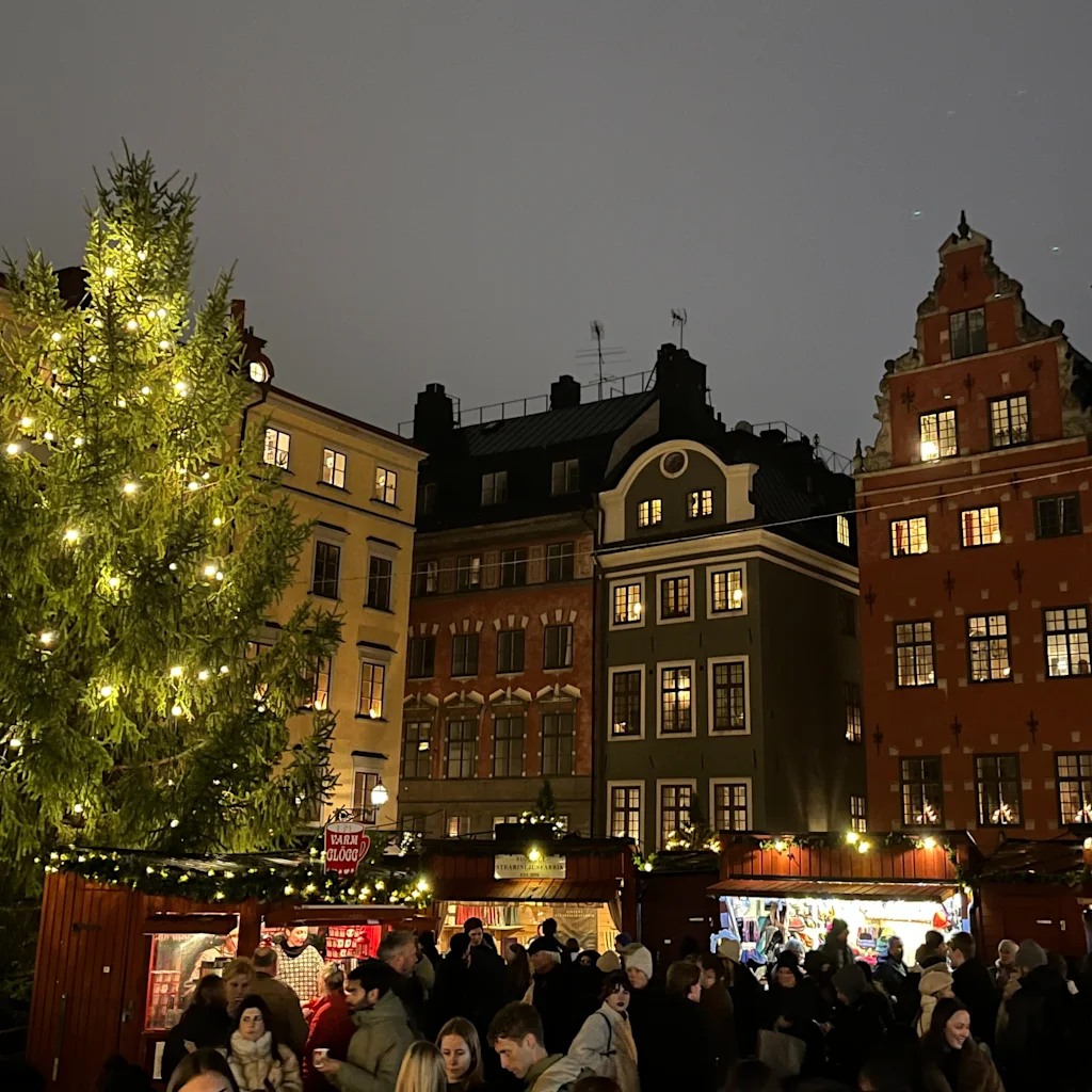 Folk samles på et hyggeligt julemarked på et historisk torv om aftenen med et lysende juletræ.