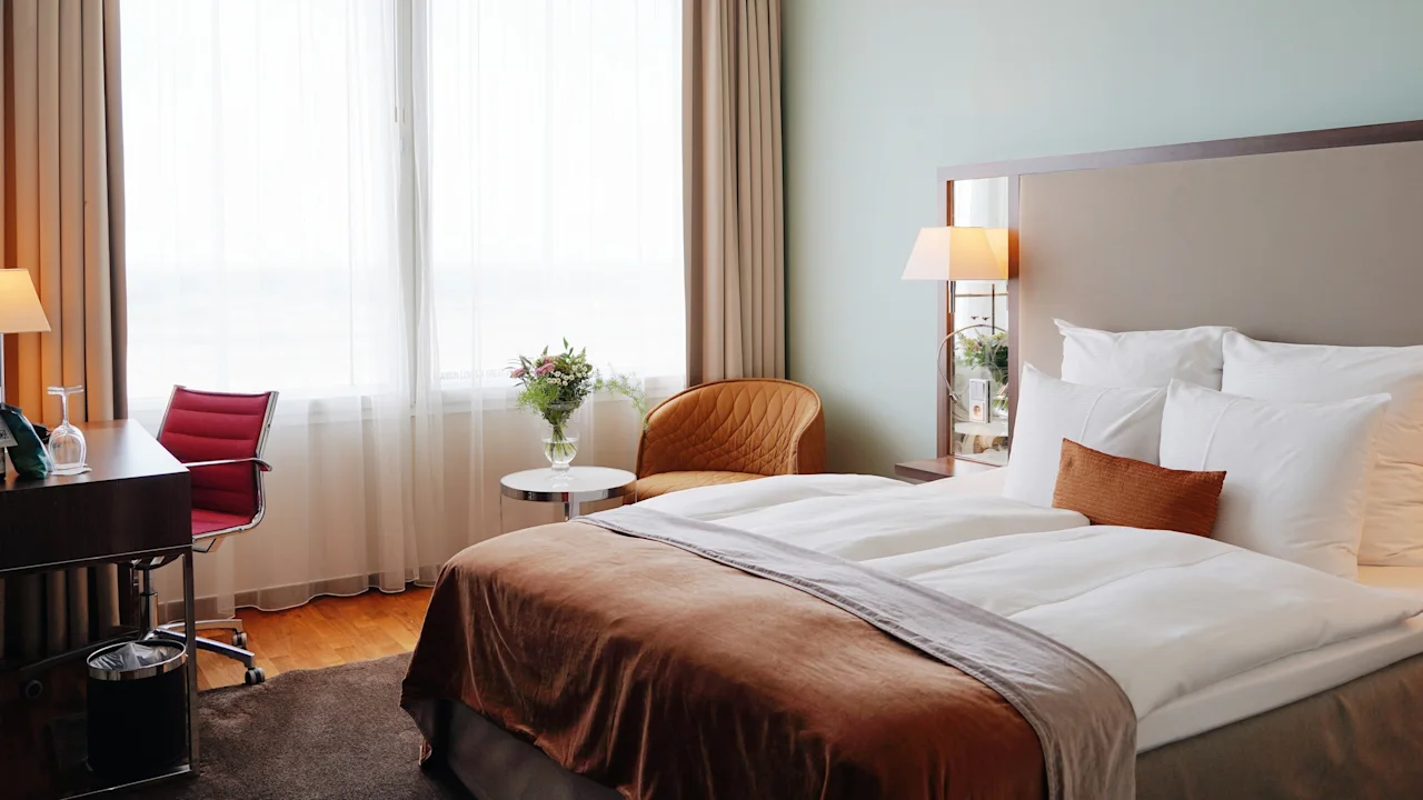 Et koselig hotellrom med en komfortabel seng, et skrivebord og et vindu. Blomster gir et friskt preg.