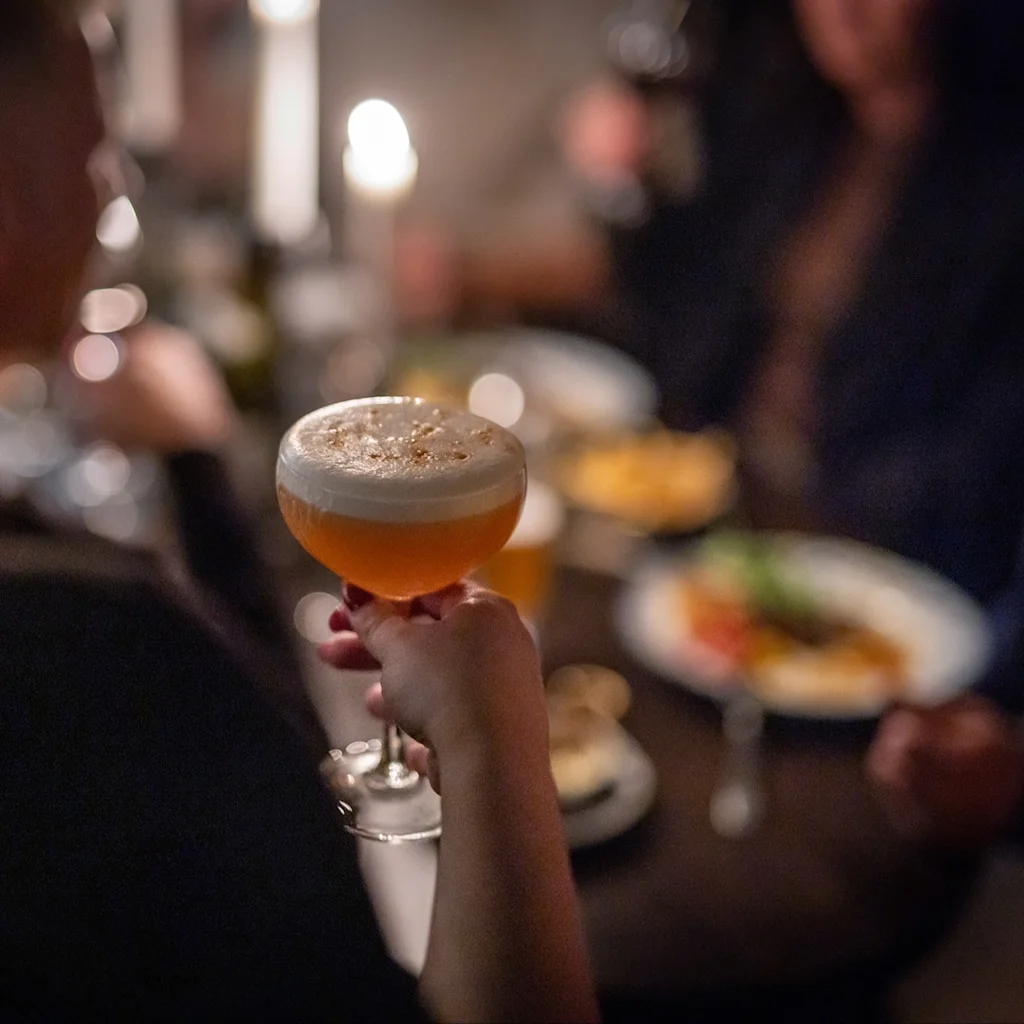 En person håller en cocktail med skummig topp i en dämpad social miljö, med andra människor och mat i bakgrunden.