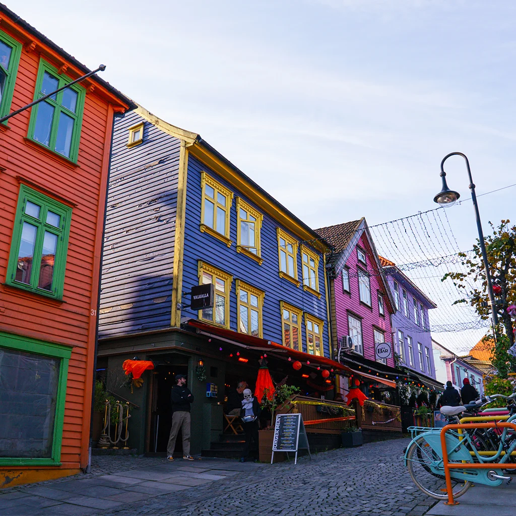 Vilkas katunäkymä värikkäine puutaloineen, käveleviä ihmisiä ja pysäköityjä polkupyöriä. Kyltissä lukee MAGASIN.