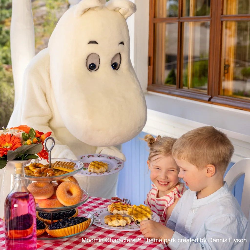 Ystävällinen hahmo tarjoilee vohveleita ja leivonnaisia kahdelle iloiselle lapselle punavalkoruudullisen pöydän ääressä.