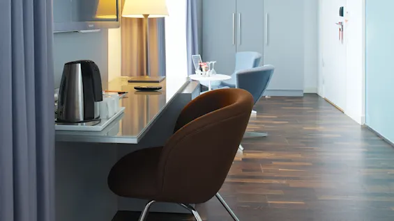 Ett modernt hotellrum innehåller en brun stol vid ett skrivbord med lampa och vattenkokare; ett fönster med gardiner och en garderob syns i bakgrunden.