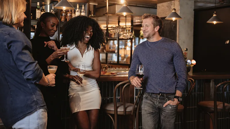 En grupp vänner njuter av drinkar och samtal på en livlig bar, med en person som skrattar glatt.