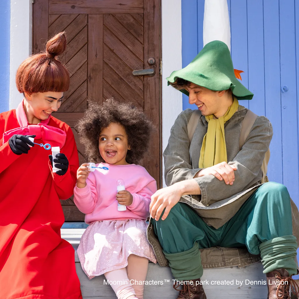 Kaksi aikuista ja lapsi puhaltavat iloisesti saippuakuplia yhdessä värikkään talon ulkopuolella, jakaen onnellisen hetken.