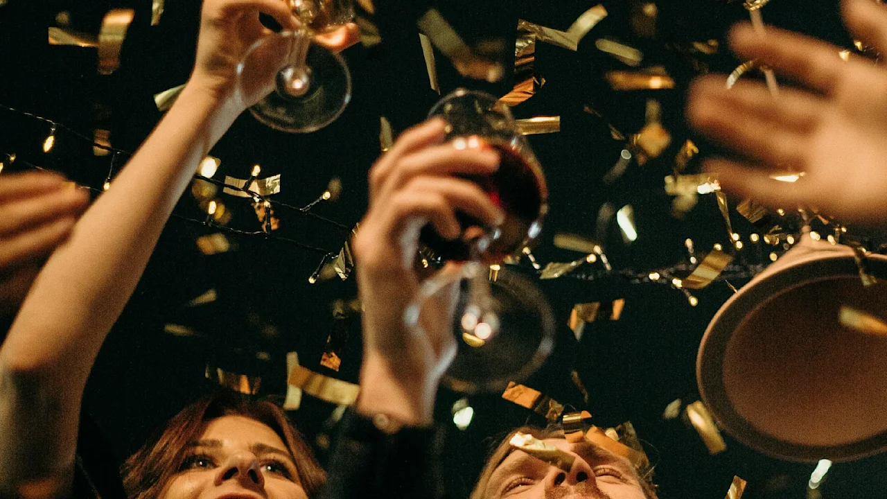 People raise glasses in celebration, surrounded by golden confetti, under dim lighting.