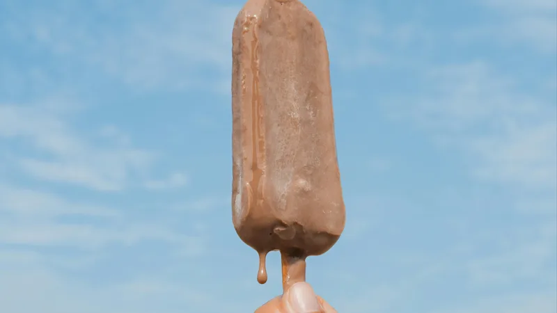 En hand håller en smältande chokladglass i bakgrunden av en blå himmel och avlägsen havshorisont.