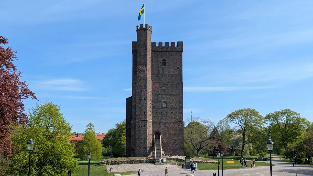 Kärnan i Helsingborg. Et historisk mursteinstårn med svensk flagg, omgitt av grønt parkområde og folk som nyter en solrik dag.