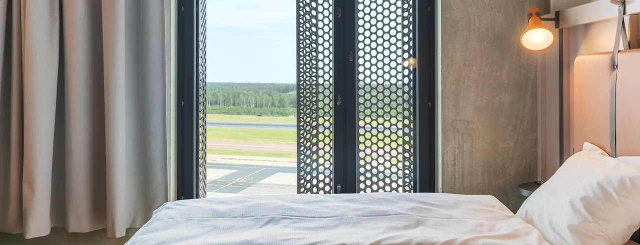 Et komfortabelt soverom med en seng, et vindu med en perforert skjerm og utsikt over en flyplass og fjerne trær.