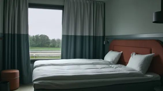 Et koselig hotellrom med en komfortabel seng, moderne innredning og et stort vindu med utsikt over et grønt landskap og vei.