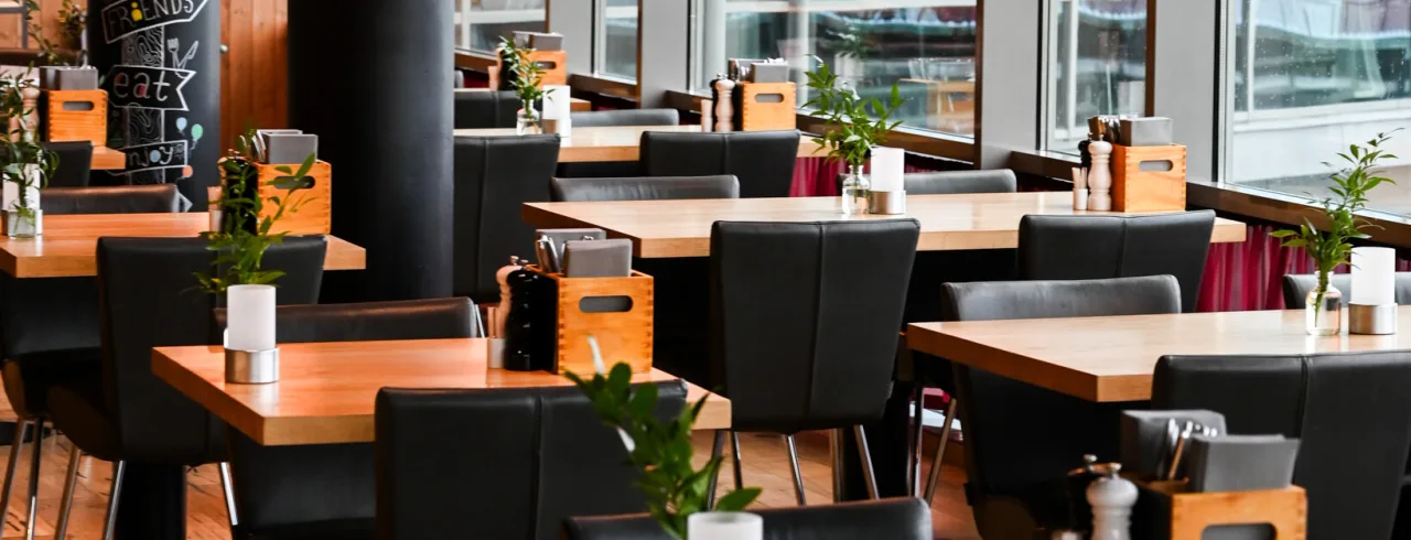 Ett modernt restauranginteriör med träbord, svarta stolar, hängande lampor och stora fönster som ger naturligt ljus.