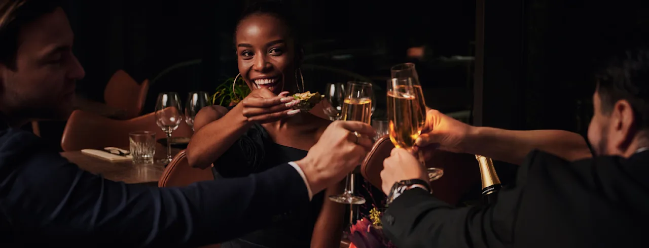 Three people toasting with champagne glasses in a dimly lit restaurant. A joyful woman holds a slice of bread. A floral arrangement decorates the table set with plates and napkins.