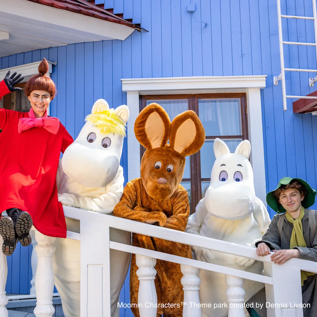 Ihmiset ja maskotit poseeraavat iloisesti kuistilla sinisen talon edessä, yksi henkilö heiluttaa ja toinen nojaa kaiteeseen.