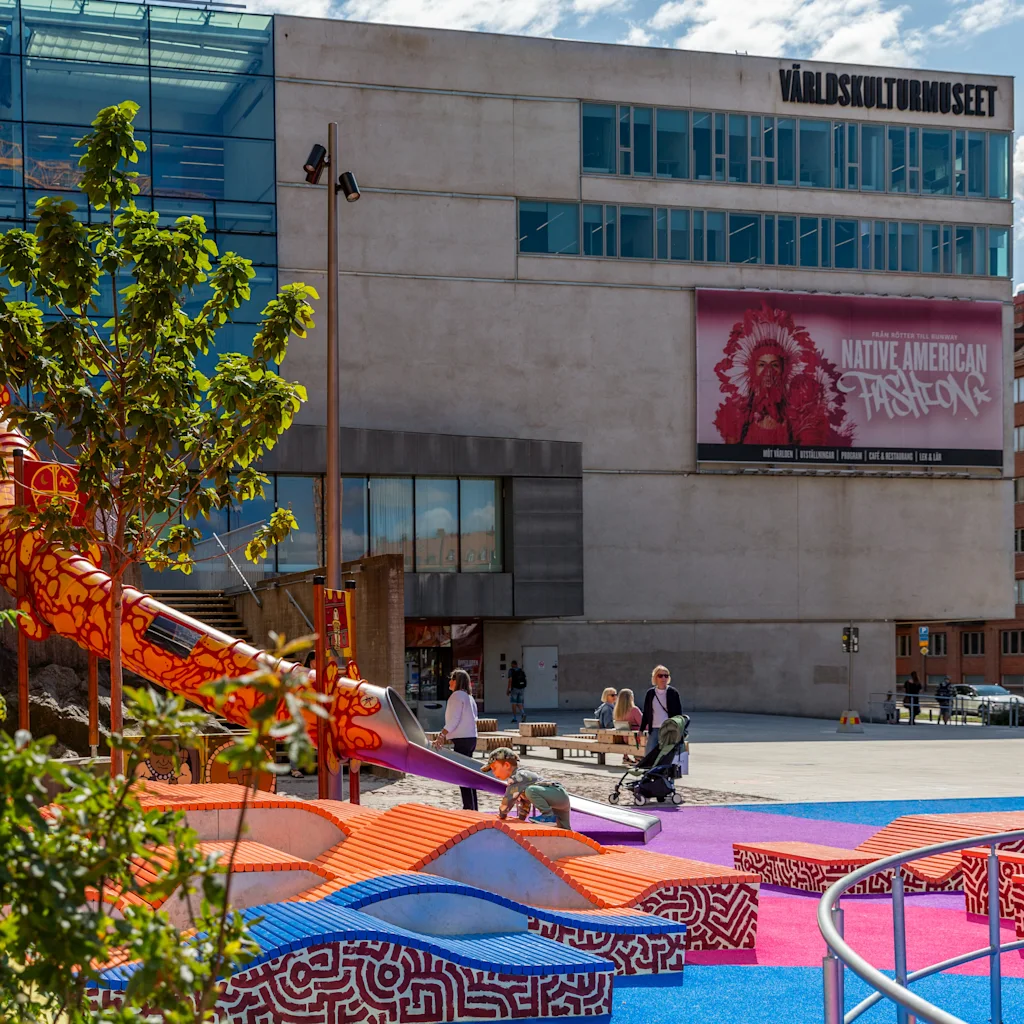 En livlig bylegeplads med en stor orange rutsjebane foran et moderne museum. Folk nyder uderummet.