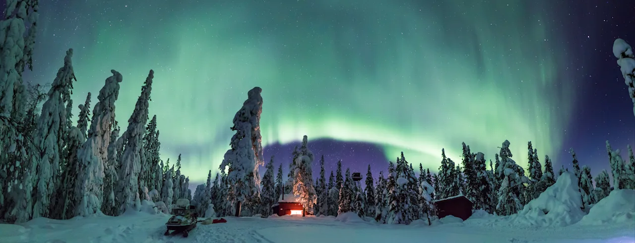 Eloisat vihreät revontulet valaisevat lumipeitteisen metsän, jossa on viihtyisä mökki ja moottorikelkka tähtitaivaan alla.