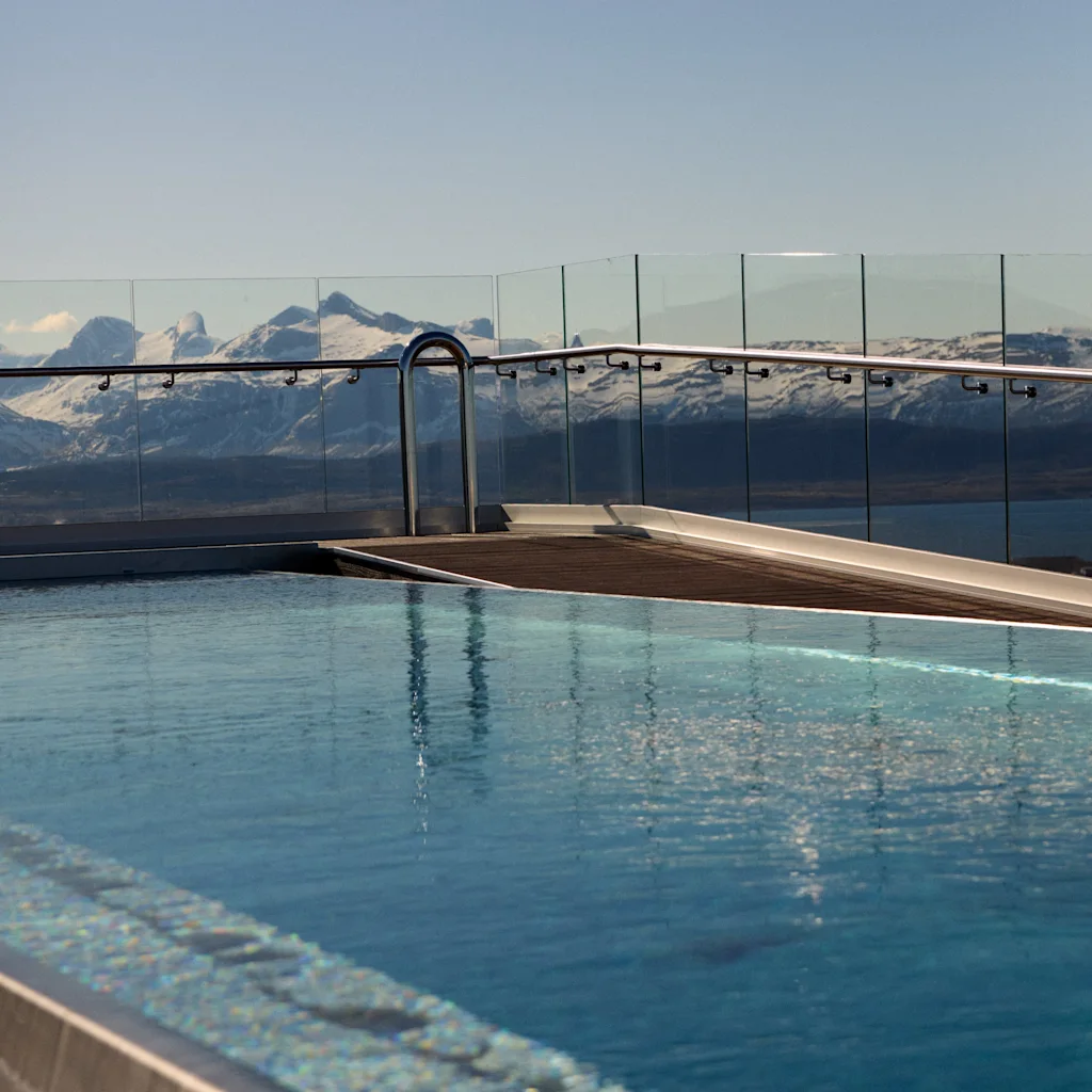A rooftop swimming pool overlooks a serene mountain range with snow-capped peaks, surrounded by clear glass railings under a blue sky.