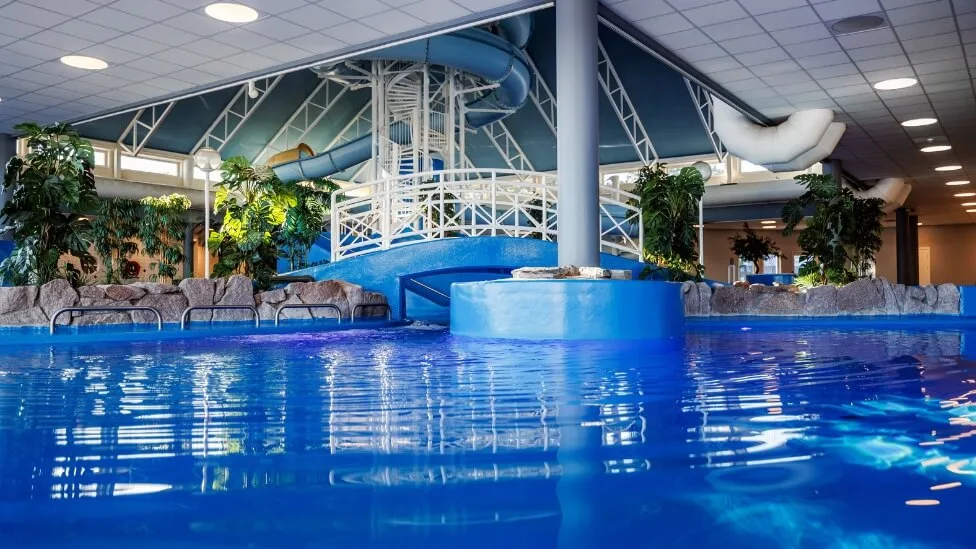 En innendørs sklie som ender i et rolig svømmebasseng; omkranset av tropiske planter og sitteområder.