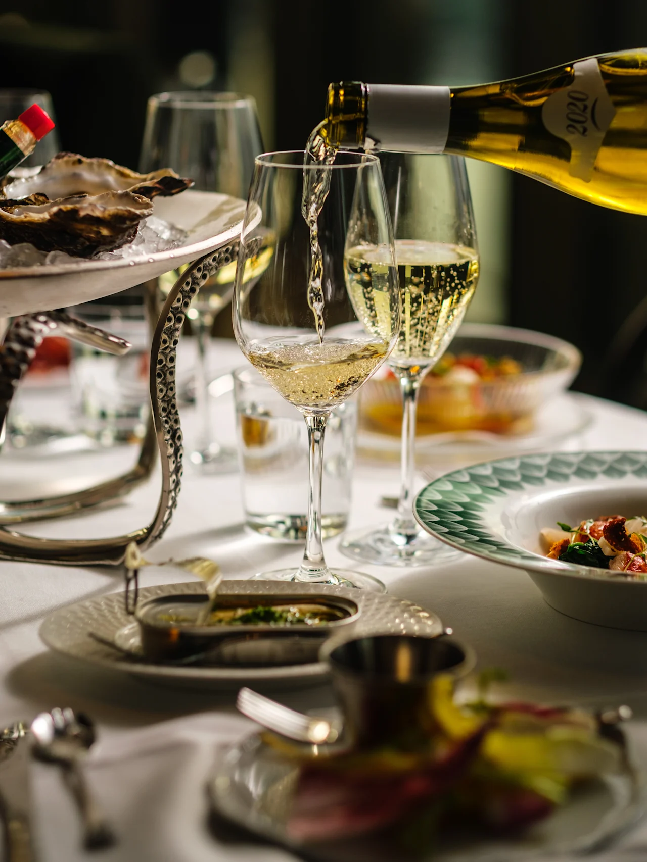 A wine bottle pours into a glass on a dining table set with oysters, glasses, and a dish of food. Atmosphere suggests fine dining.