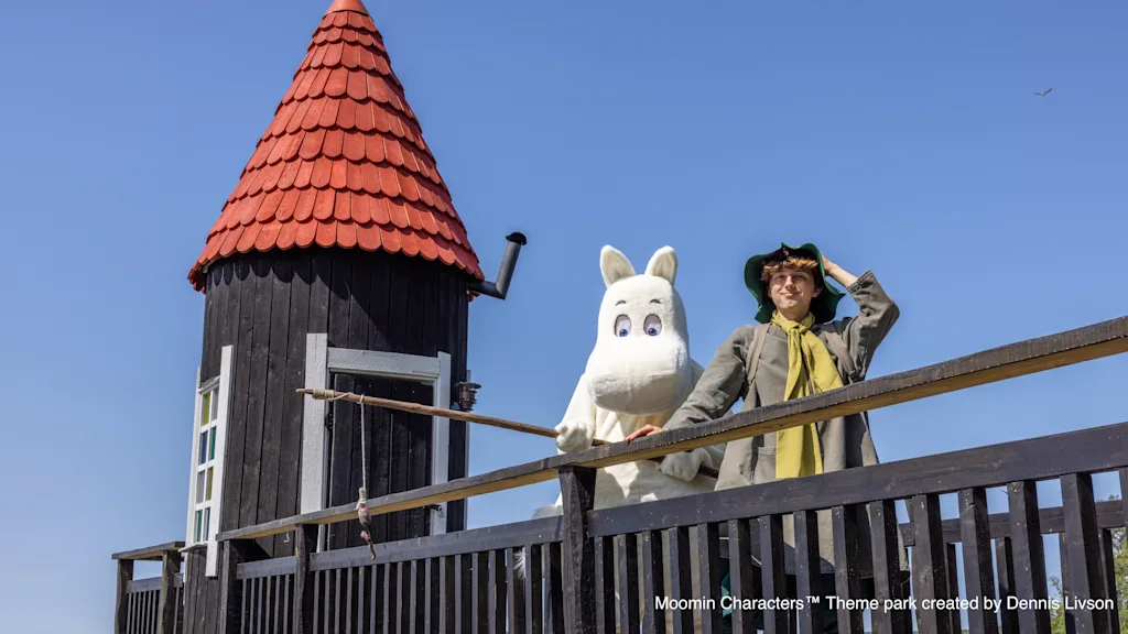 Henkilö ja Muumihahmo puulaiturilla mustan tornin ja punaisen katon vieressä aurinkoisena päivänä.