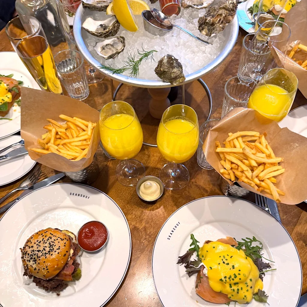 Ett bord dukat för en måltid med SKANSHOF-tallrikar, med hamburgare, eggs benedict, pommes frites, ostron och drycker.