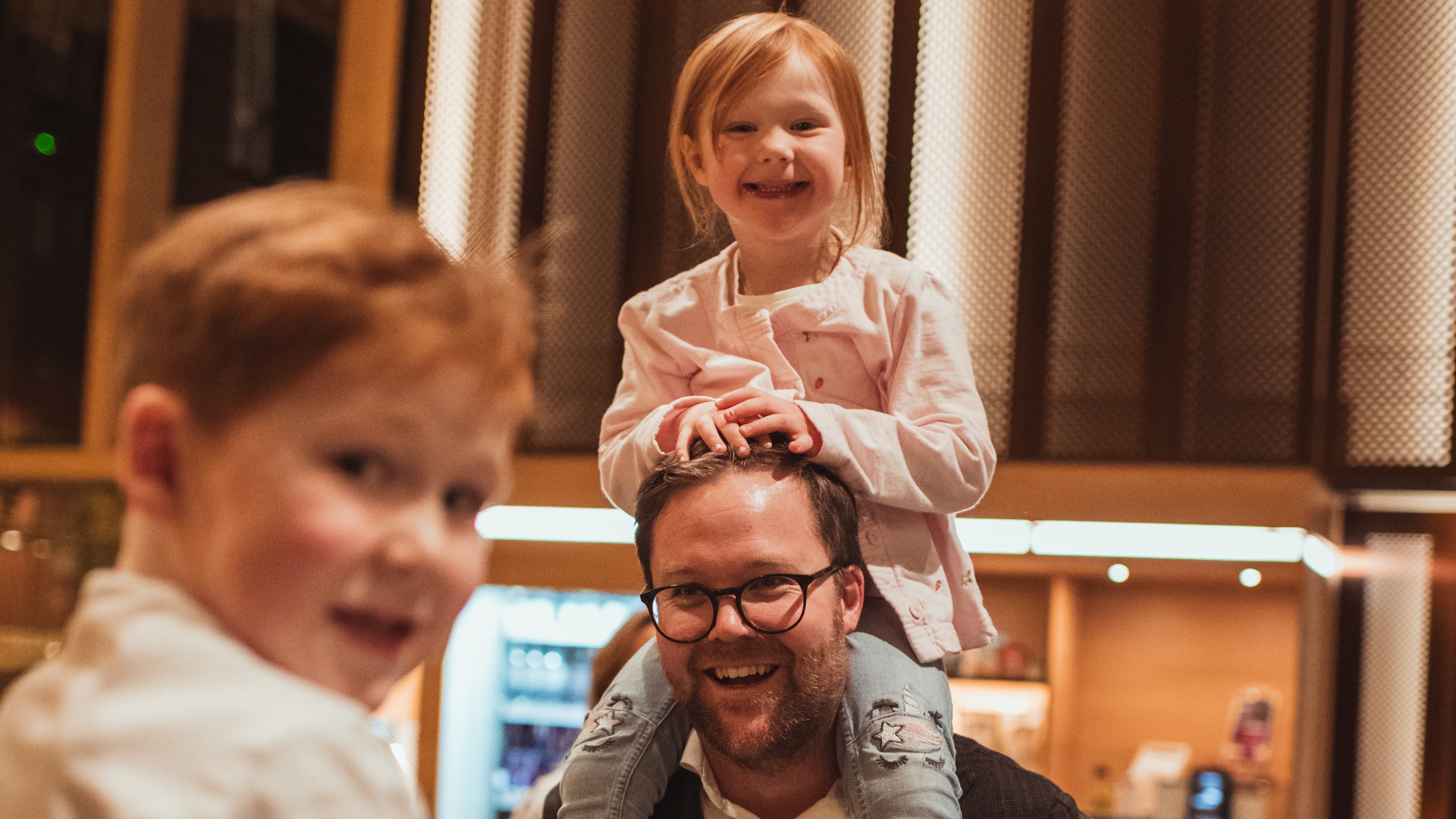 En person bærer et barn på skuldrene, begge smiler lyst. Et annet barn smiler i forgrunnen, og fanger et lykkelig øyeblikk.