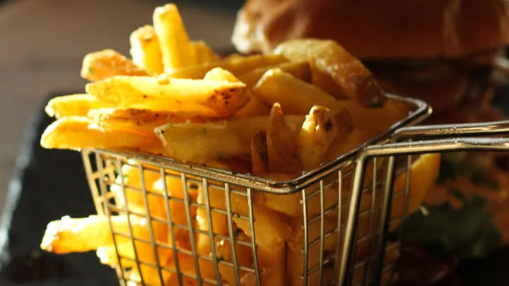 Gyllene pommes frites i en metallkorg med en hamburgare i bakgrunden, på ett bord.
