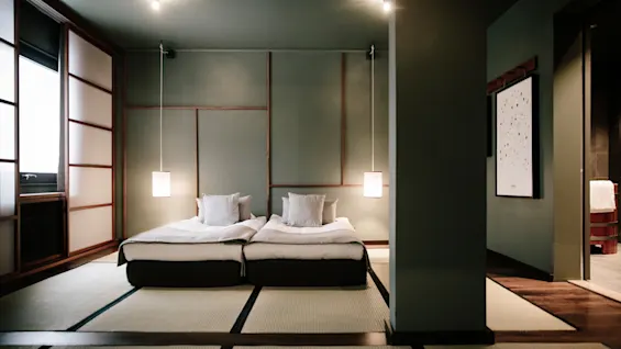 To senger med hvitt sengetøy i et rom med grønne vegger, tre-detaljer og hengende lamper; en rolig, moderne estetikk.