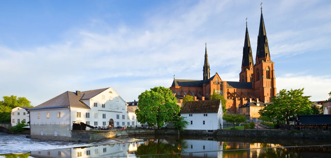 A scenic view of Uppsala with a river reflecting historic buildings and a grand cathedral under a blue sky.