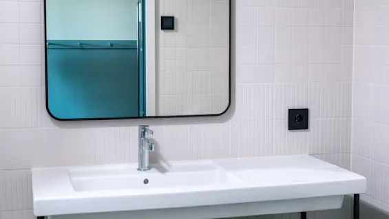 Mirror reflecting a partially open bathroom door; hangs above a white sink with a metal stand. A towel dangles from the stand. Context includes beige tiled walls and a round light fixture.