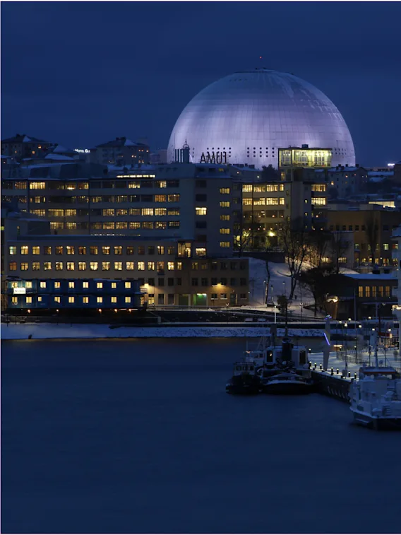 Stadsvy på natten med en stor upplyst kupol, upplysta byggnader och båtar vid en snötäckt kaj. Text: AMOI.